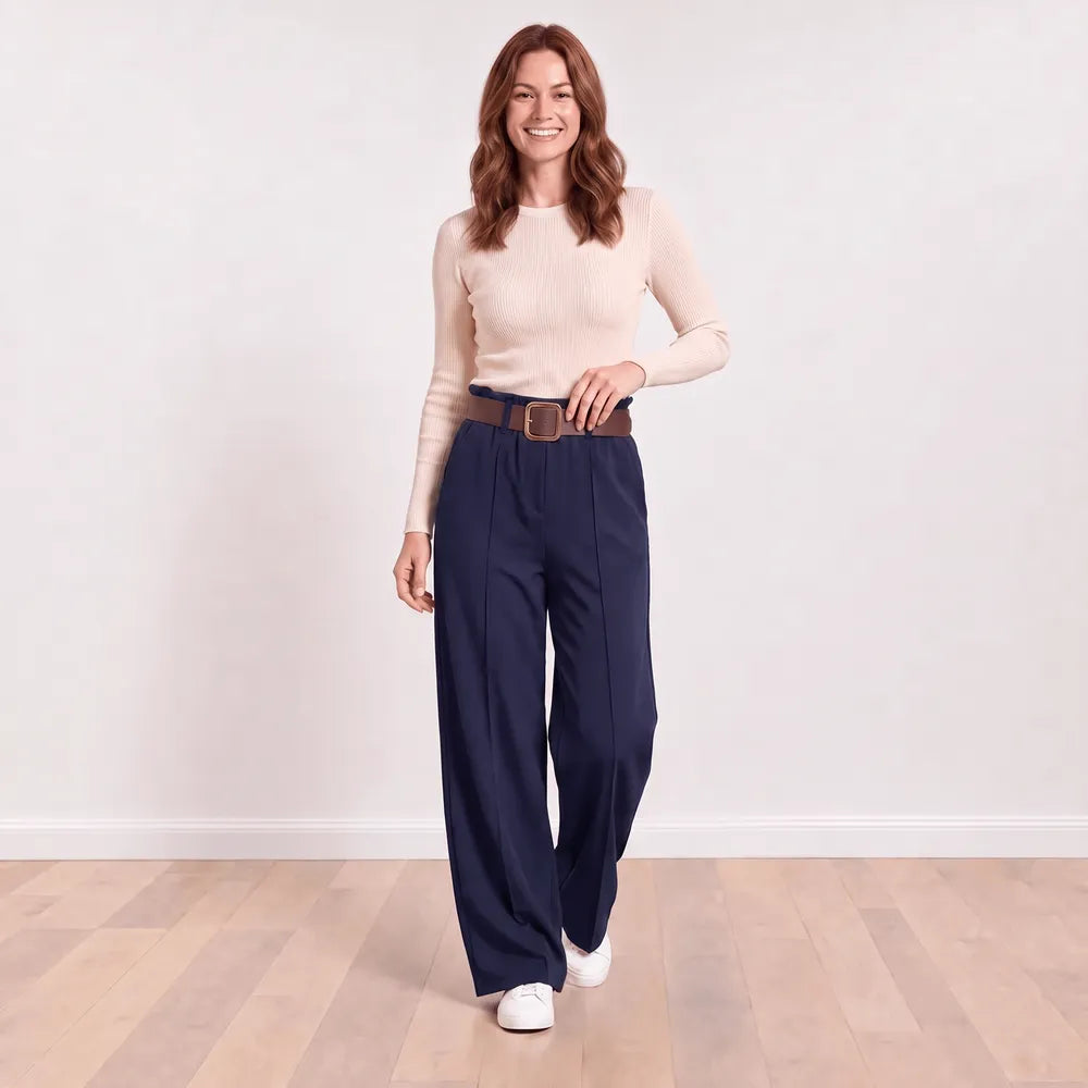 Woman wearing navy Ellana wide-leg paperbag-waist trousers with a cream ribbed knit top, brown leather belt and white trainers, front view with hand on belt
