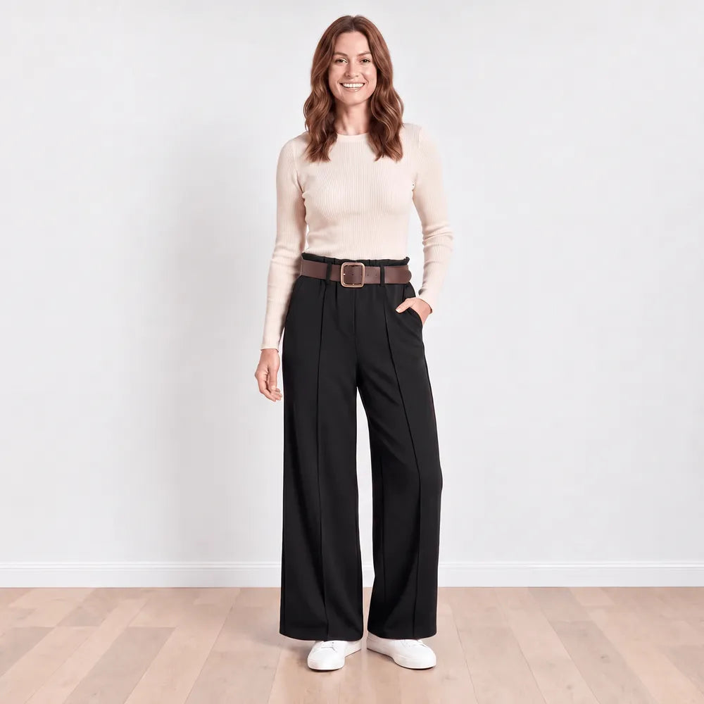 Model wearing Ellana wide-leg pleated trousers in black with a cream ribbed knit top, brown leather belt and white trainers, front view with hand in pocket